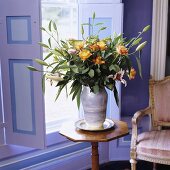 A bunch of flowers featuring yellow roses on a delicate wooden occasional table in front of lilac-painted window shutters