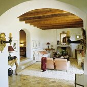 A view through a arched doorway into an open living room in a finca with a rustic wood beam ceiling and a white sofa in front of a fireplace
