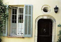 Bars in front of a window with grey wooden shutters and a dark wooden front door with a stone facade and a round window above the door in a light yellow facade