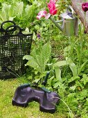 Knee pad and black plastic hand tote in front of a flower bed with watering can hanging from a tree
