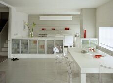 Modern, white open plan kitchen diner with glass fronted cupboard units and table with perspex chairs.