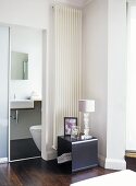 A modern, minimalist bedroom decorated in neutral colour, view through sliding door to ensuite bathroom, washbasin, toilet, wooden floor, wall mounted radiator, table, lamp,