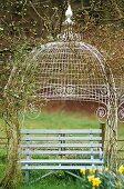 Wooden bench seat under an ornate wrought iron canopy with climbing plants