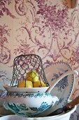 Drahtkorb über Zitronen auf Keramiksieb und Schalenstapel vor Tapete mit Blumen-Ornamentmuster
