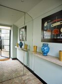 A modern hallway with mirrored wall with reflection of open door, brick floor, shelf, blue vase, abstract painting, sense of space,