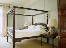 A traditional bedroom with painted panelling, carved wooden four poster bed, bedside table, lamps