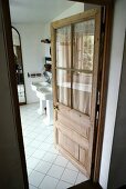 Halbverglaste, offene Holztür in traditionellem Bad mit weissen, diagonal verlegten Fliesen und Säulenwaschbecken