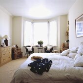 Eine schwarz-weiße Jacke liegt auf dem Bett mit weißer Bettwäsche, das zusammen mit ein Paar Korbtruhen in einem weißen Schlafzimmer steht