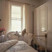 weiße Vorhänge umgeben das Fenster eines weißen Schlafzimmers, das mit einem Eisenbett und einen großen Wandschrank eingerichtet ist