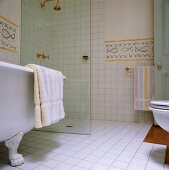 Eine freistehende Badewanne mit Klauen-Füßen steht vor der modernen Nasszelle mit einer Glaswand des weißgefliesten Badezimmers