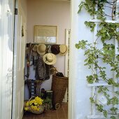 Garderobe mit Hüten und Jacken, zu sehen durch eine offene Haustür, daneben eine Kletterpflanze mit Spalier
