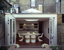 Open conservatory doors leading to a terrace and a view of a dining table with white bucket chairs in an open-plan kitchen