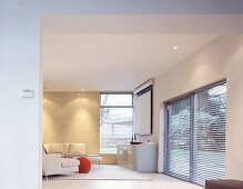 Blick durch breiten Durchgang auf offener Designer Wohnnraum mit weißem Sofa und Sideboard neben Fenster mit halbgeschlossener Jalousie
