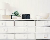 weiße Schubladenkommode mit einer Sammlung weisser Schalen und zwei Zimmerpflanzen obenauf