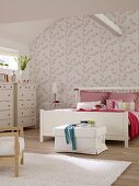 Weisses Doppelbett und Kommode im Schlafzimmer mit rosa-weiß geblümter Tapete und Dachbalken