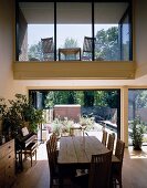 Rustikaler Esstisch mit Stühlen vor offener Terrassentür und Blick auf verglasten Ausschnitt mit Loggia im Obergeschoss
