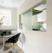 A black plastic, retro-style bucket chair next to a dining table with a large, modern serving hatch with a view into the kitchen