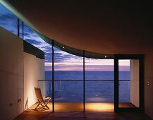 Chair on illuminated terrace beyond empty living room with view of evening sky under a floating roof curving upwards at one side