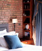 Corner of bedside table with bookcase and cushions and curtain in blue and beige fabric in front of brick wall