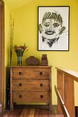 Abstract portrait hanging on a yellow wall above chest of drawers