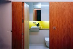 Blick durch offene Tür aus Holz in kleines designtes Bad mit Waschbecken und gelb lackierter Rückwand