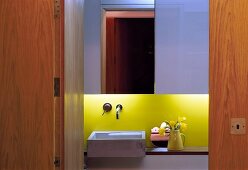 Blick durch offene Tür aus Holz in designtes Bad mit Waschbecken und gelb lackierter Rückwand mit indirekter Beleuchtung