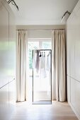 Dressing room with white fitted wardrobes and clothing on coat hangers on metal frame in front of French windows