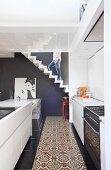 White, open-plan, designer kitchen-dining room with strip of mosaic tiles in traditional pattern and man walking up sculptural, zigzag stairs