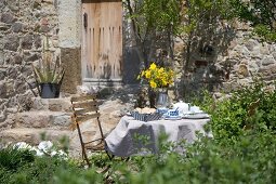 Gedeckter Frühstückstisch im Garten mit gelbem Blumenstrauss vor mediterranem Natursteinhaus