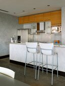 Bar stools in front of counter and concrete walls in open-plan kitchen with glass, wood and stainless steel fronts