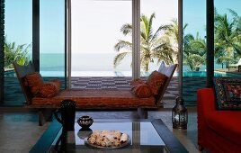 Wooden recamier with loose cushions in front of large windows with view of wooden terrace and palms behind infinity pool