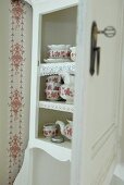 Weisses Küchenbuffet mit offener Tür und Blick auf traditionelles Service mit rot-weißem Blumenmuster