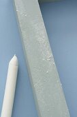 White candle next to grey wooden batten on pastel blue background