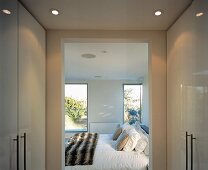 Ankleideraum mit weiss glänzenden Einbauschränken und offenem Durchgang zu freundlichem Schlafzimmer