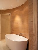 Freistehende Badewanne vor holzverkleideter Wand im ovalen, modernen Bad