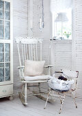 Weiß gestrichene Zimmerecke eines Holzhäuschens mit altem Schaukelstuhl und Kissen neben verrostetem Metallstuhl