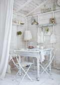 Weiß gestrichene Zimmerecke eines Holzhäuschens mit weißem Küchentisch und schlichten Gartenstühlen