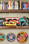 Books and tin toys on wooden shelves and painted plates hung on the wall below
