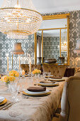 Chandelier above festively set table with tablecloth and upholstered chairs in front of mirror on wall with patterned wallpaper