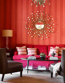 Corner of living room with red striped wallpaper and decorative, sun-shaped mirror above deep pink sofa