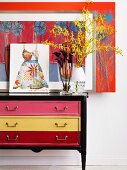 Blumenvase und Bilder auf Vintage Kommode mit verschieden farbigen Schubladen vor der Wand