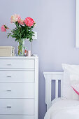 Vase of flowers on white chest of drawers next to bed against lilac wall