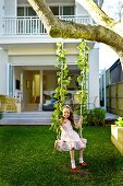 Girl sitting on swing hanging from tree in front of modern house