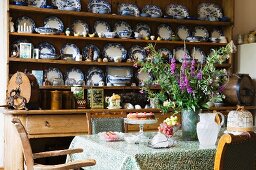 Kostbare Sammlung blauweisser Teller im Holzregal und Küchentisch mit antiken Stühlen im Vordergrund