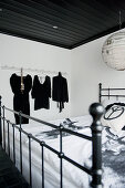 Antique-style metal bed below black-painted wooden ceiling and Japanese paper lampshade; black clothing on hangers in background