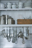 Crockery, stainless steel jugs and cooking utensils hanging from hooks on white shelves