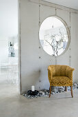 Bed of pebbles and vintage velvet armchair in front of exposed concrete partition wall with aperture