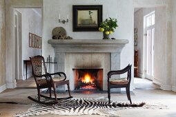 Vintage rocking chair and antique armchair on zebra-skin rug in front of open fire in simple house