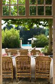 View through open terrace doors of wicker chairs at a set table with pool in Mediterranean garden in background