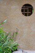 Wand im Garten mit Blick durch eine runde Öffnung auf geschnürte Bambusstäbe